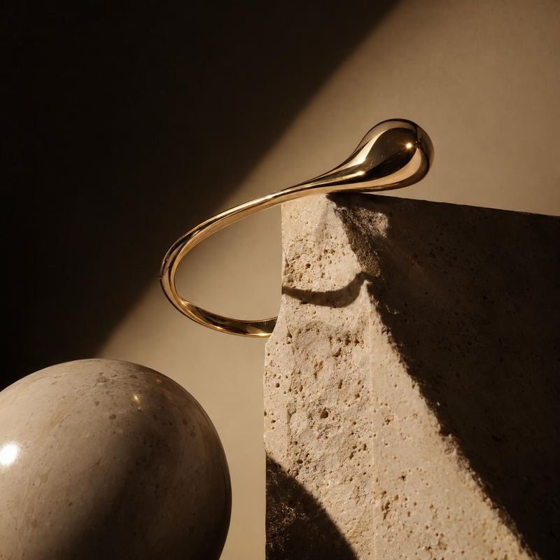 gold cuff bracelet jewelry product photo: A polished gold cuff curves over a rough stone block beside a rounded stone form, with warm directional light creatin…