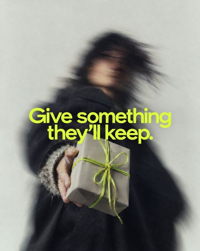 wrapped gift box action & motion product photo: A motion-blurred woman in a dark knit outfit holds a simple gift box tied with vivid lime-green ribbon against…