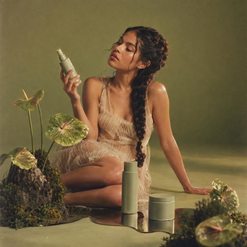 skincare spray and cream product in hand product photo: A woman in a sheer neutral dress admires a skincare spray amid matching green containers and leafy plan…