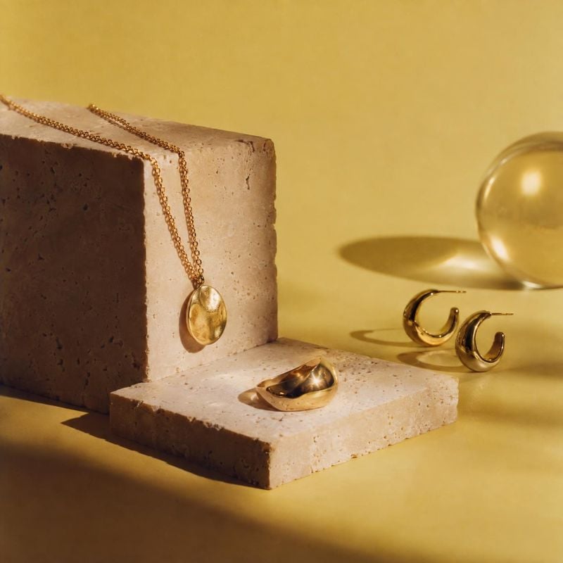 gold jewelry set jewelry product photo: A gold necklace, earrings and ring are artfully arranged on pale stone blocks against a warm yellow backdrop, with dire…