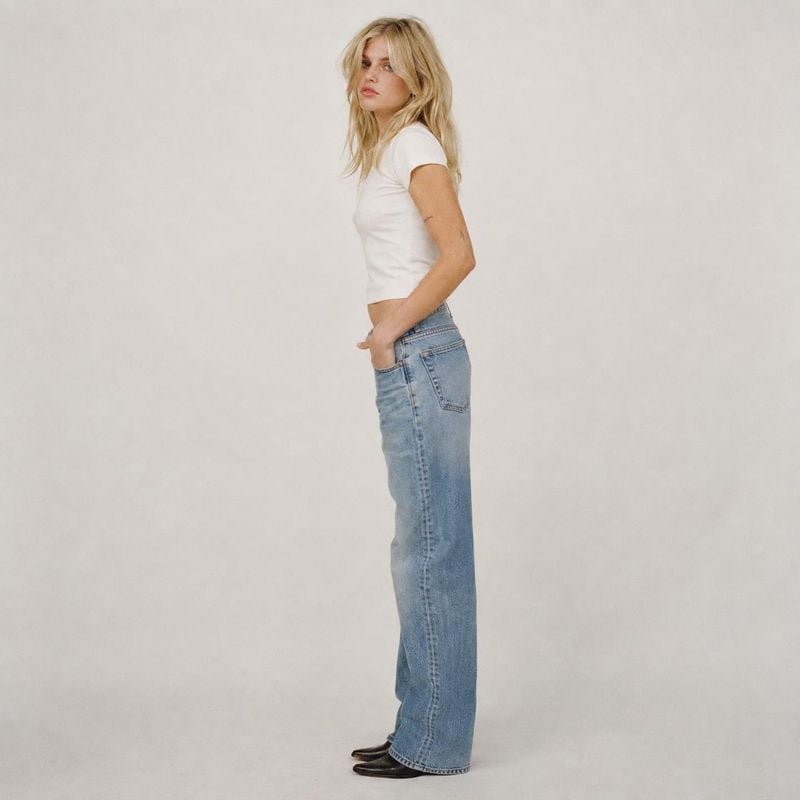 blue jeans on-body detail product photo: A blonde model poses in a white fitted T-shirt, loose blue jeans and dark shoes against a softly lit neutral studio ba…