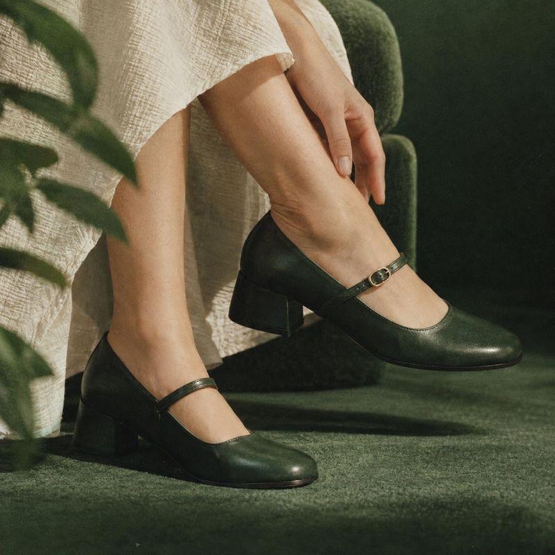 black Mary Jane shoes on-body detail product photo: A model in a light textured skirt wears black Mary Jane shoes against deep green upholstery and carpeting,…