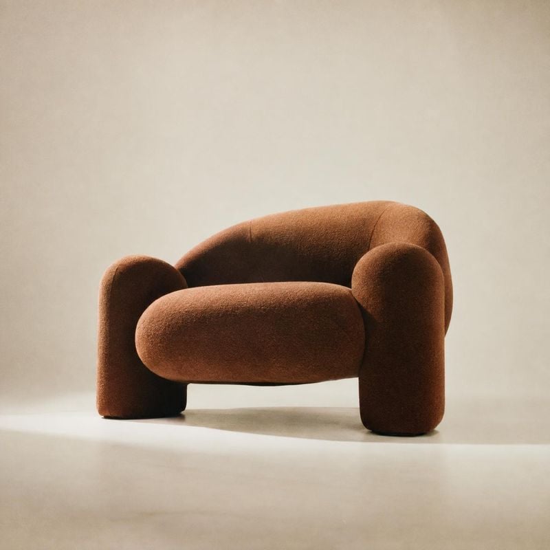 upholstered lounge chair home product photo: A sculptural rust-brown upholstered lounge chair sits alone against a warm, seamless beige studio backdrop, softly…