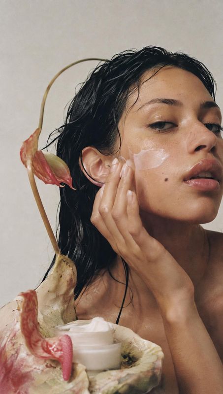 face cream beauty close-up product photo: A woman smooths cream onto her cheek beside an open jar nestled among sculptural botanical forms, photographed in sof…