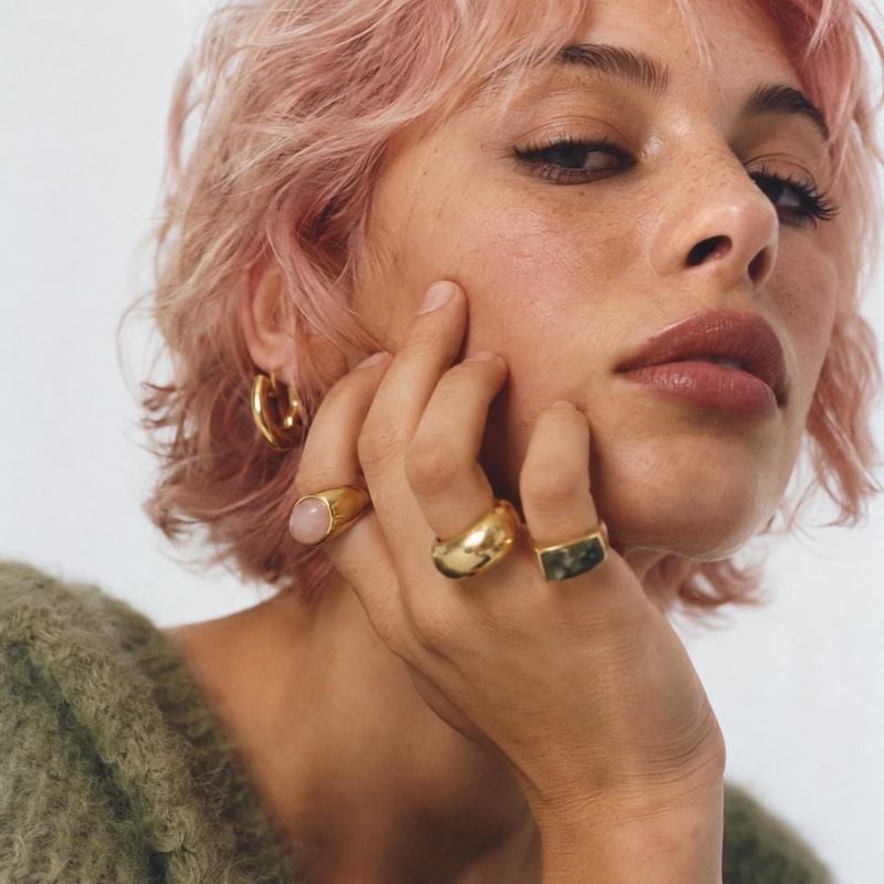 gold rings and hoop earrings beauty close-up product photo: A close-up portrait of a woman with tousled pink hair, wearing sculptural gold rings and hoop earri…