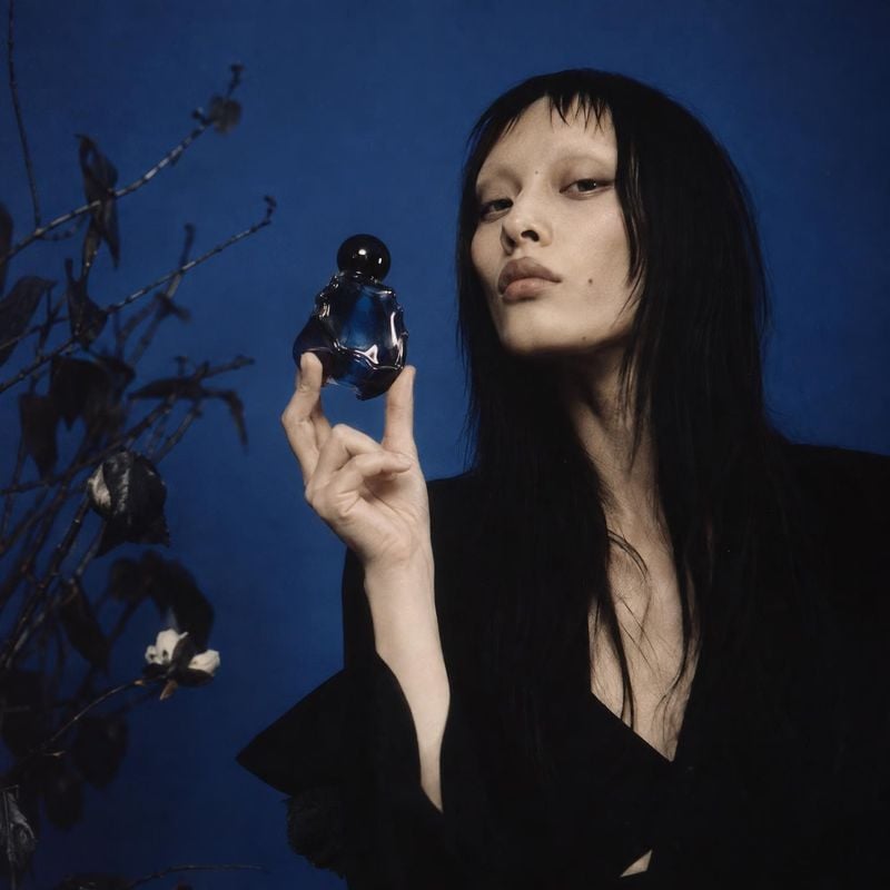 perfume beauty close-up product photo: A dark-haired model in black holds a small blue perfume bottle against a deep blue studio backdrop with delicate branche…