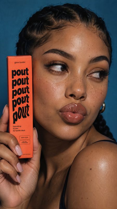 lip tint beauty close-up product photo: A woman with glossy lips and softly defined eye makeup holds a coral lip-tint box against a deep blue studio backdrop,…