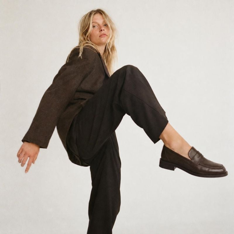 leather loafers model try-on product photo: A blonde model poses dynamically against a softly lit off-white studio backdrop, wearing a dark tailored outfit and…