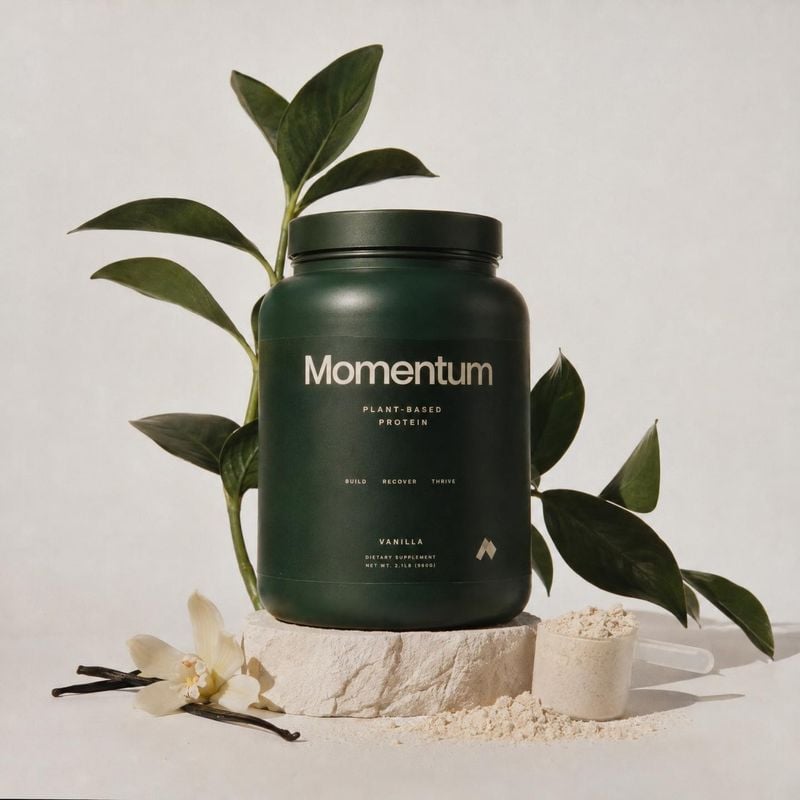 plant-based protein powder personal care product photo: A dark green tub of vanilla plant-based protein powder sits on a pale stone block among leafy branches,…