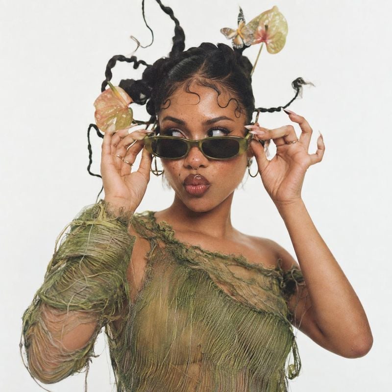 sunglasses model try-on product photo: A model in sculptural olive-toned styling adjusts narrow sunglasses against a plain white studio backdrop, with soft eve…