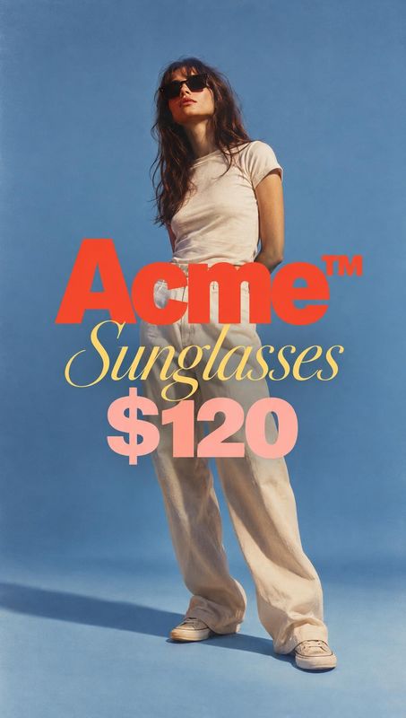 sunglasses jewelry product photo: A model in sunglasses, a white tee, and loose cream trousers poses against a blue studio backdrop beneath bold red, yellow, a…