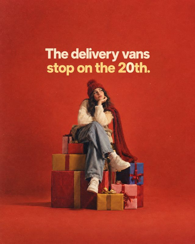 gift boxes christmas product photo: A woman in a red knit hat, cream sweater and jeans sits among wrapped gift boxes against a warm red studio backdrop, beneat…