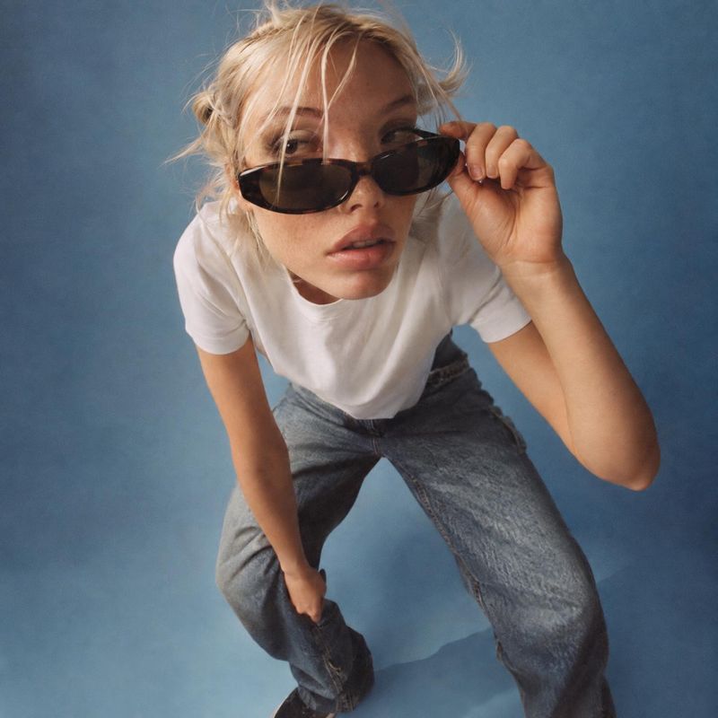sunglasses on-body detail product photo: A blonde model leans toward the camera in a white T-shirt and jeans while adjusting dark sunglasses against a softly l…
