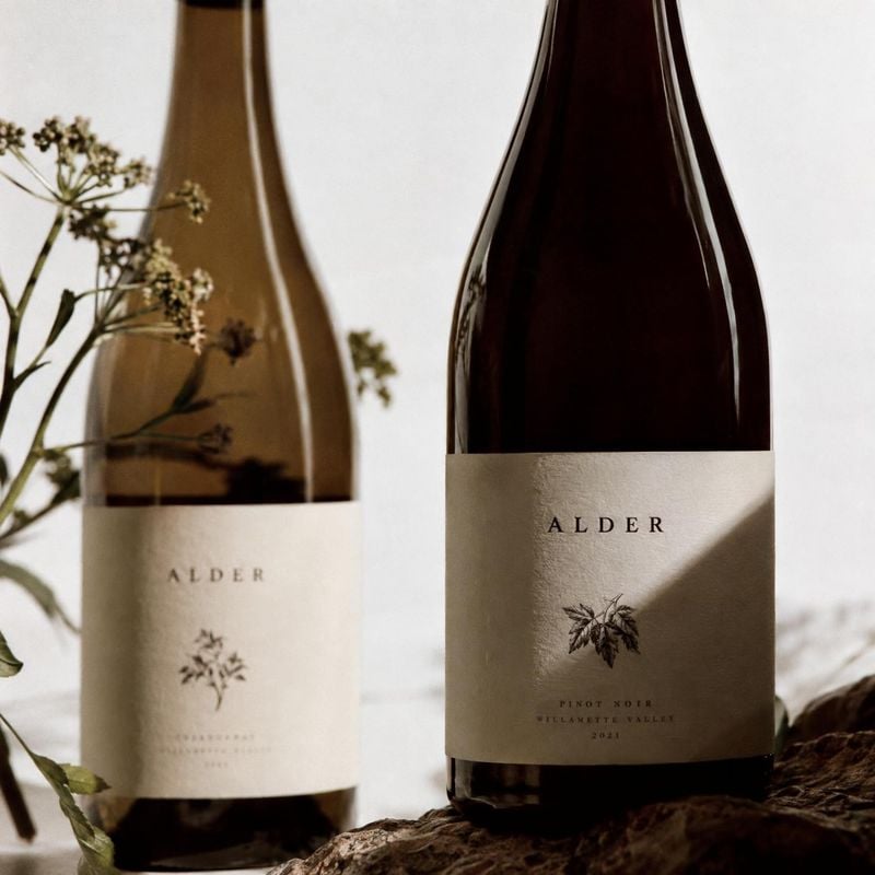 wine bottles close-up detail product photo: Two dark wine bottles with cream labels stand among delicate dried botanicals and natural textures, softly lit agai…