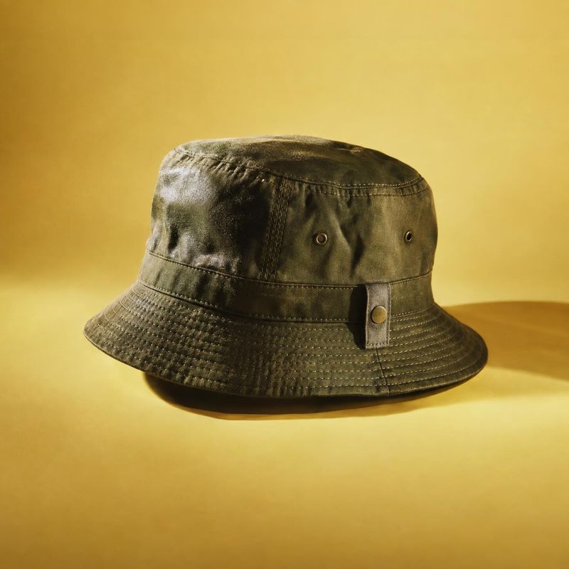 olive bucket hat fashion product photo: An olive bucket hat is displayed against a warm yellow seamless backdrop, with directional light emphasizing its textur…
