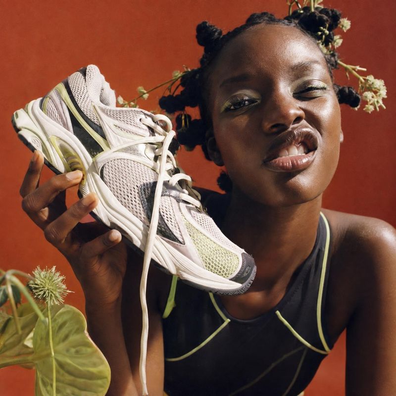 running shoe product in hand product photo: A woman in a black athletic top holds a pale mesh running shoe beside her face against a warm terracotta backdrop,…