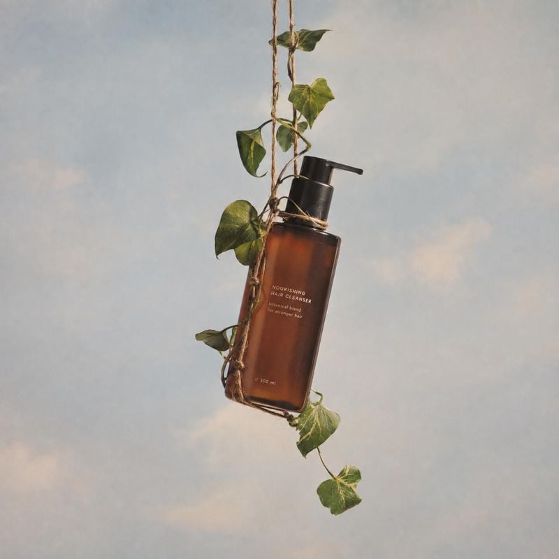 hair cleanser floating product product photo: An amber hair cleanser bottle hangs among trailing green vines against a softly clouded pale backdrop, with warm…