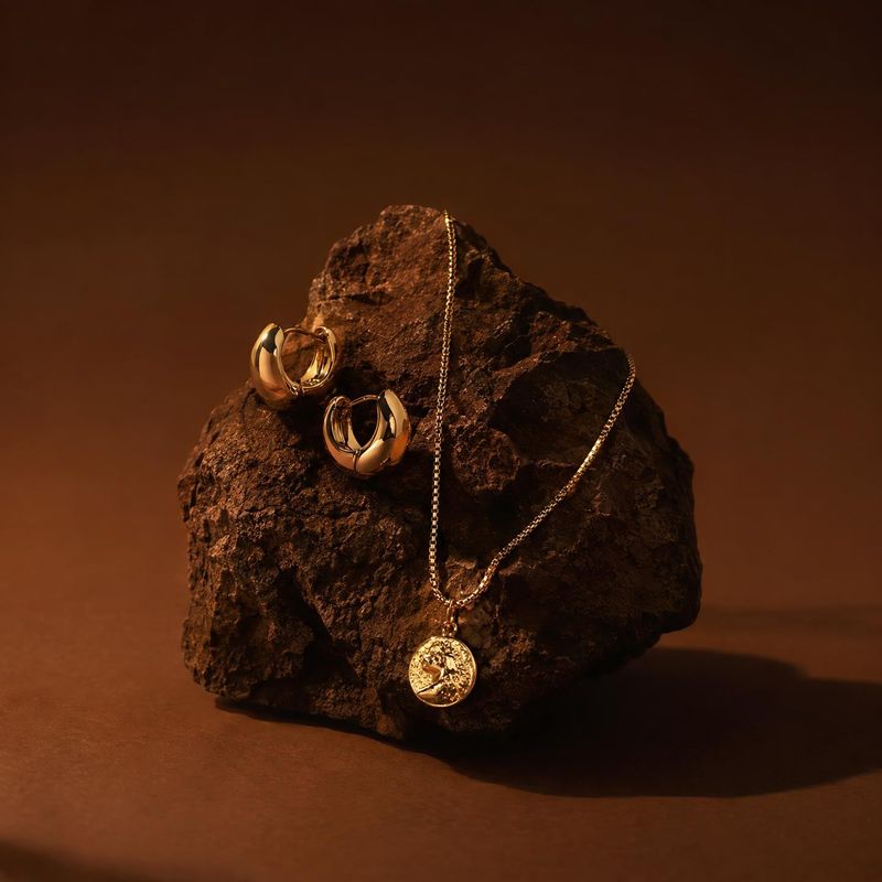 gold necklace and earrings black friday product photo: A gold necklace and sculptural hoop earrings are arranged on a rugged dark rock against a warm brown stu…