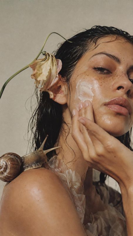 face cream beauty close-up product photo: A close, softly lit beauty portrait shows a woman smoothing cream onto her cheek, with a dried flower and snail addin…