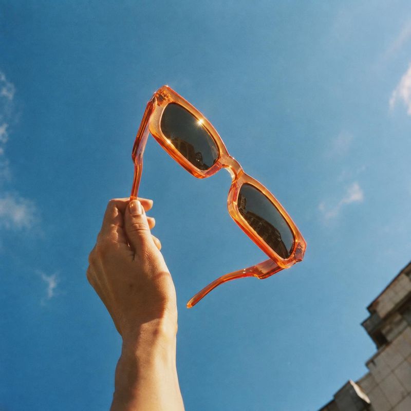 sunglasses product in hand product photo: A hand lifts translucent amber sunglasses against a vivid blue sky, with warm sunlight and a city building at the edg…