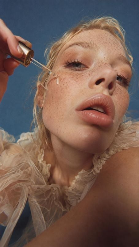 facial serum beauty close-up product photo: A close-up beauty portrait shows a freckled blonde woman applying a drop of serum with a glass dropper against a de…