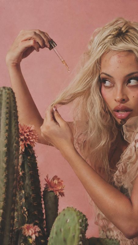 hair oil beauty close-up product photo: A blonde model applies hair oil with a dropper against a soft pink backdrop, with flowering cacti adding a botanical to…