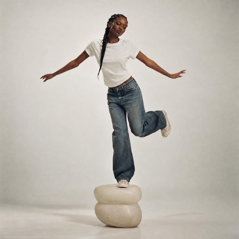 blue jeans action & motion product photo: A woman in a white T-shirt, relaxed blue jeans, and light sneakers balances playfully on a sculptural stone stool aga…