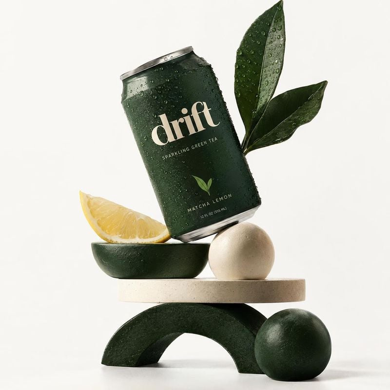 sparkling green tea food and drink product photo: A chilled dark-green can of sparkling matcha lemon tea is staged with a lemon wedge, glossy leaves, and sculp…