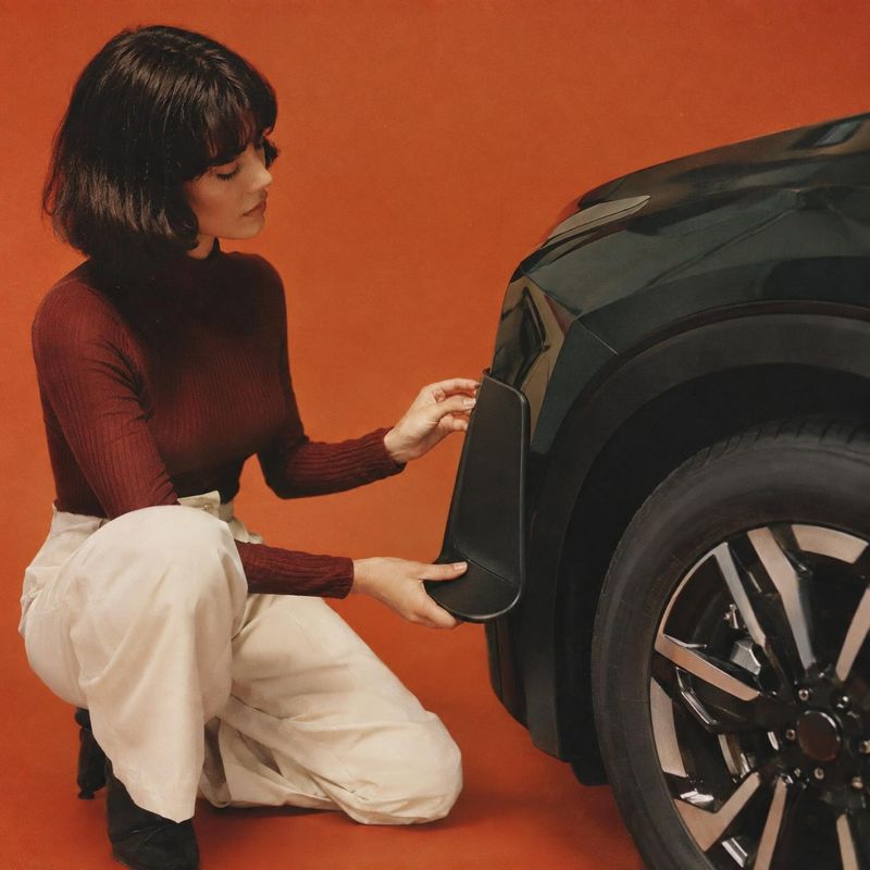 automotive splash guard close-up detail product photo: A woman in a burgundy top and cream trousers fits a black splash guard beside a vehicle’s wheel against…