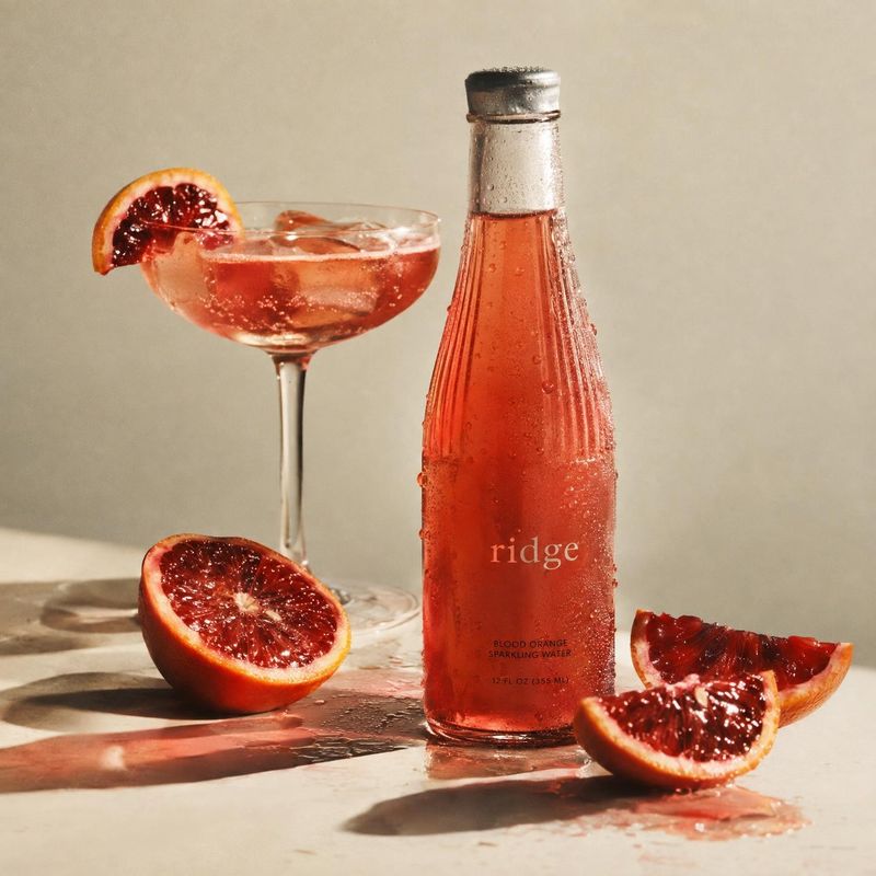 blood orange sparkling water food and drink product photo: A chilled bottle of blood orange sparkling water stands with a citrus-garnished cocktail and sliced…