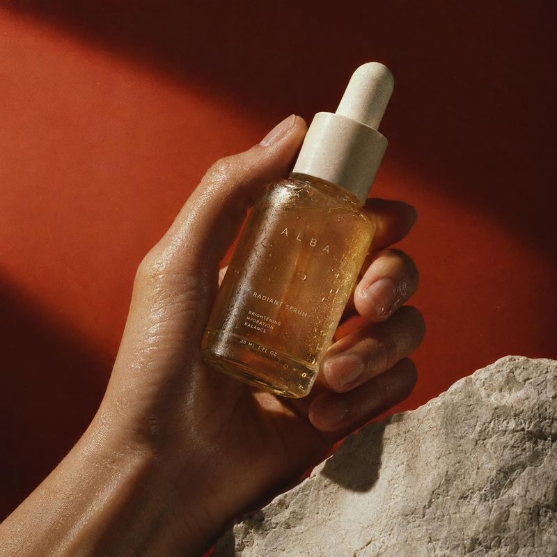 radiant serum product in hand product photo: A hand presents a golden dropper bottle of radiant serum against a warm terracotta backdrop, with textured stone i…