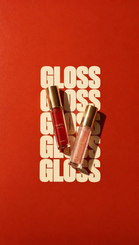 lip gloss flat lay product photo: Two lip gloss tubes with metallic caps are arranged diagonally over repeated cream lettering on a vivid red background, with…