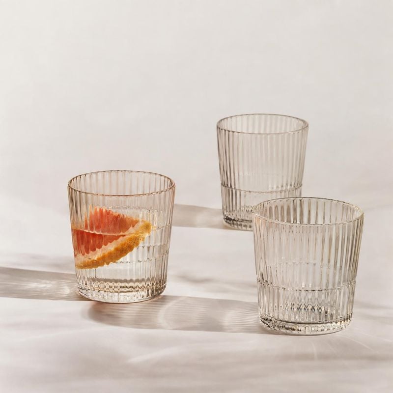 ribbed glass tumblers group lineup product photo: Three clear ribbed tumblers are arranged on a softly lit neutral surface, with a citrus slice in one glass an…