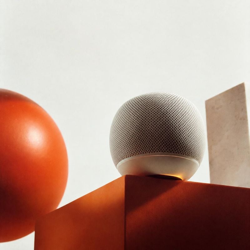 spherical smart speaker electronics product photo: A fabric-covered spherical smart speaker sits on a rust-red geometric pedestal beside an orange sphere, lit…