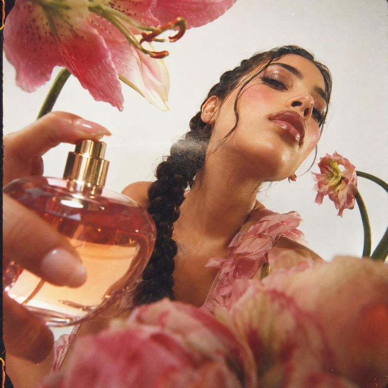 perfume beauty close-up product photo: A woman with a glossy braided hairstyle holds a pink perfume bottle among oversized pink flowers, photographed in soft b…