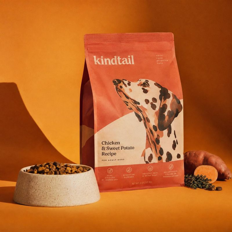 dog food pet product photo: A coral Kindtail dog-food bag stands beside a bowl of kibble and sweet potatoes on a vivid orange set, lit with warm directional li…