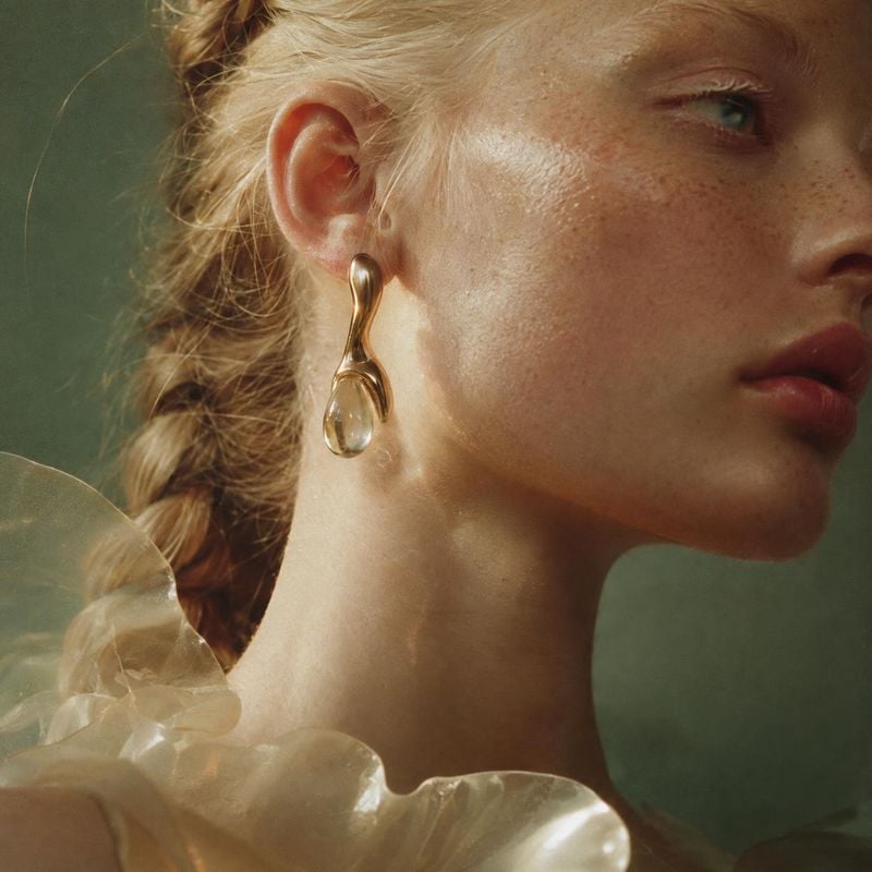 gold drop earrings on-body detail product photo: A close, softly lit portrait highlights sculptural gold drop earrings against a muted green studio backdrop, w…