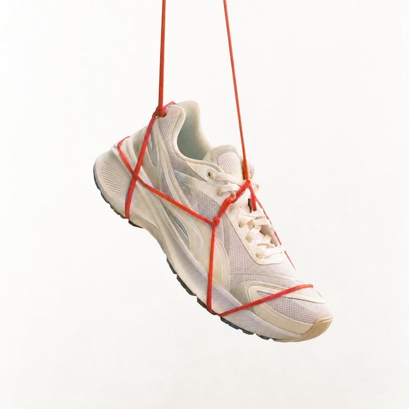 running shoe fashion product photo: A white running shoe hangs at an angle from vivid red cords against a bright, minimal studio background, creating a clean a…