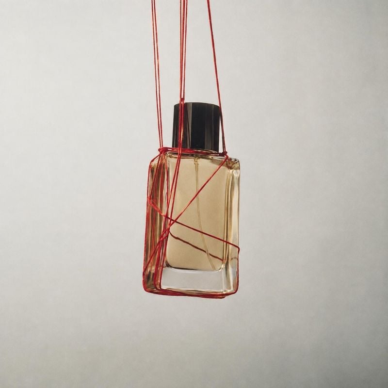 perfume bottle floating product product photo: A glass perfume bottle hangs suspended in red threads against a softly lit pale gray studio background, creating…