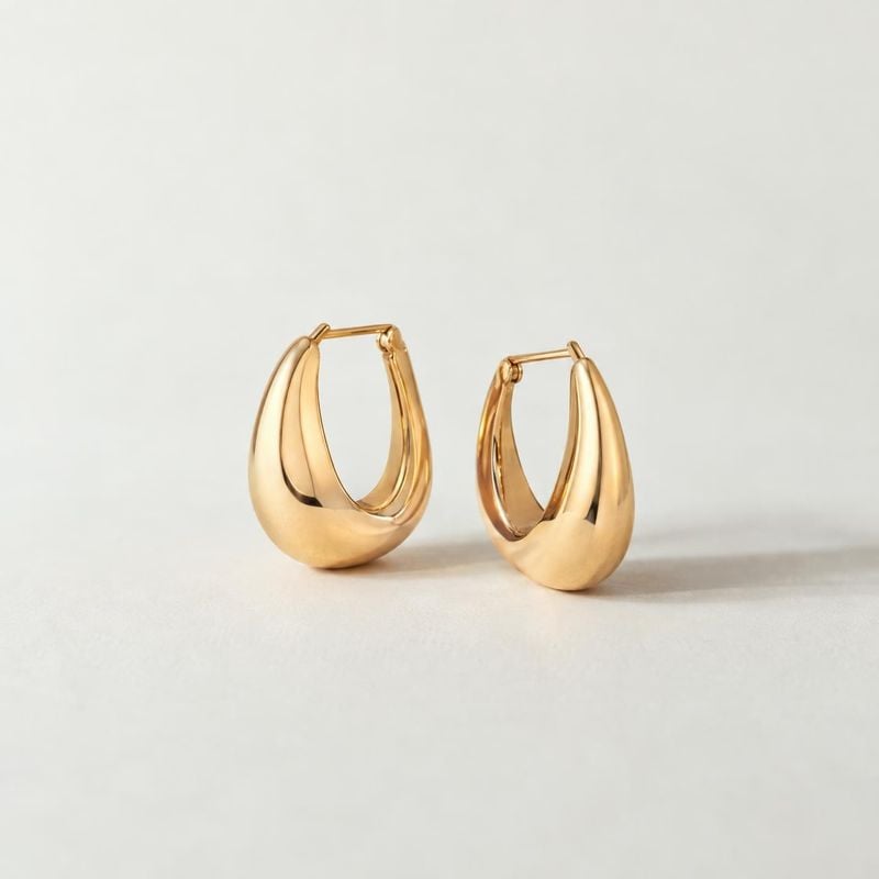 gold hoop earrings jewelry product photo: A polished pair of sculptural gold hoop earrings stands against a softly lit ivory background, with gentle shadows an…
