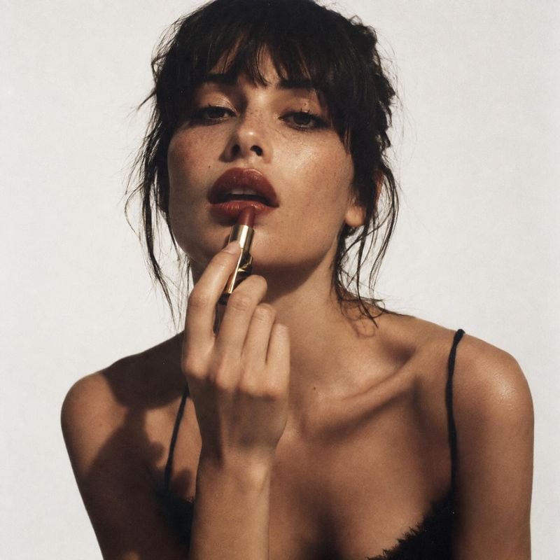 lipstick beauty close-up product photo: A woman with tousled dark hair applies warm-toned lipstick in a close beauty portrait against a softly lit off-white st…