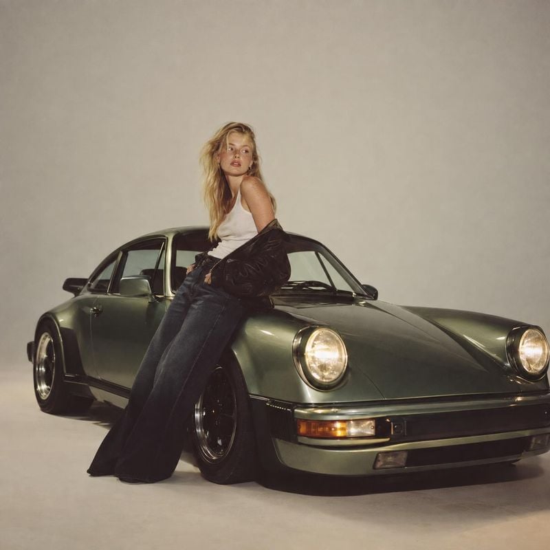 women's casual outfit vehicle exterior product photo: A blonde model in a white tank top, dark jeans and a black jacket poses against a muted studio backdrop w…