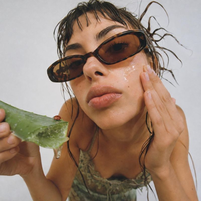 aloe vera leaf beauty close-up product photo: A close-up beauty portrait shows a woman in tinted sunglasses applying gel from a fresh aloe leaf to her cheek ag…