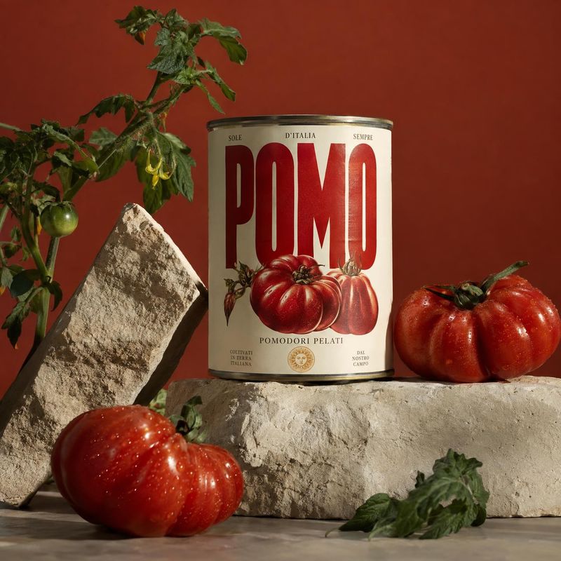 can of peeled tomatoes ingredients product photo: A can of peeled tomatoes is staged among ripe tomatoes, a tomato plant, and rough stone props against a warm…