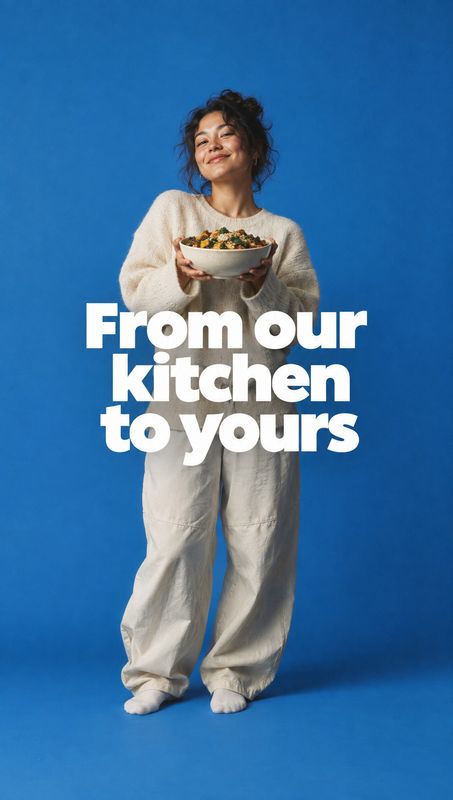 prepared meal bowl food and drink product photo: A smiling woman in relaxed cream-colored clothing holds a bowl of food against a vivid blue studio backdrop, w…