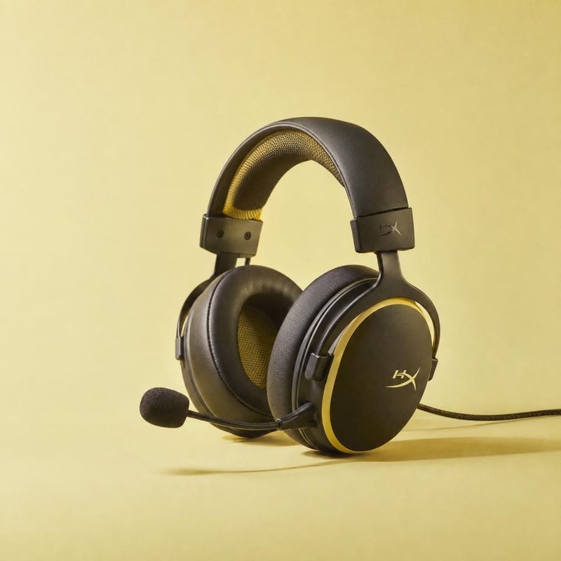 gaming headset electronics product photo: A black over-ear gaming headset with gold accents and a boom microphone is presented against a warm yellow studio bac…