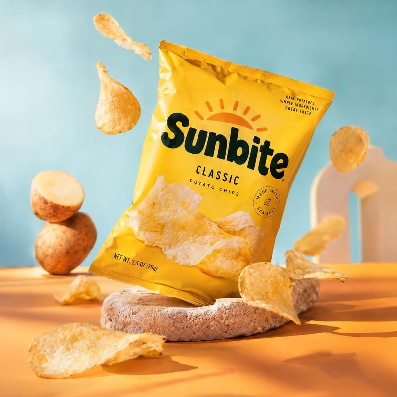 potato chips food and drink product photo: A bright yellow bag of potato chips is staged among scattered chips and potatoes on a warm orange surface against a…