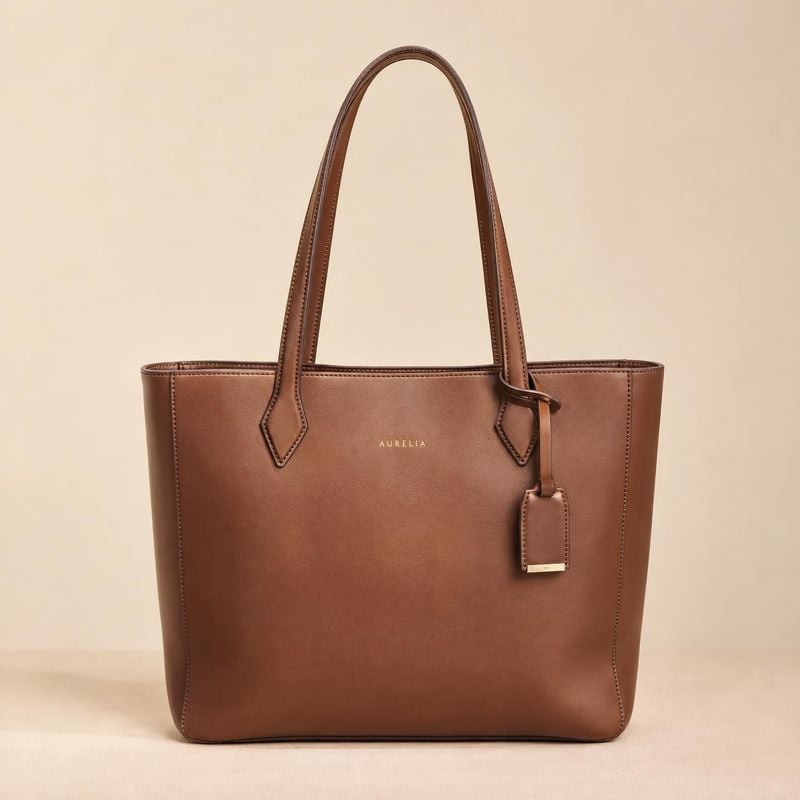 leather tote bag jewelry product photo: A structured brown leather tote with long handles and a hanging tag is centered against a warm beige studio backdrop, l…