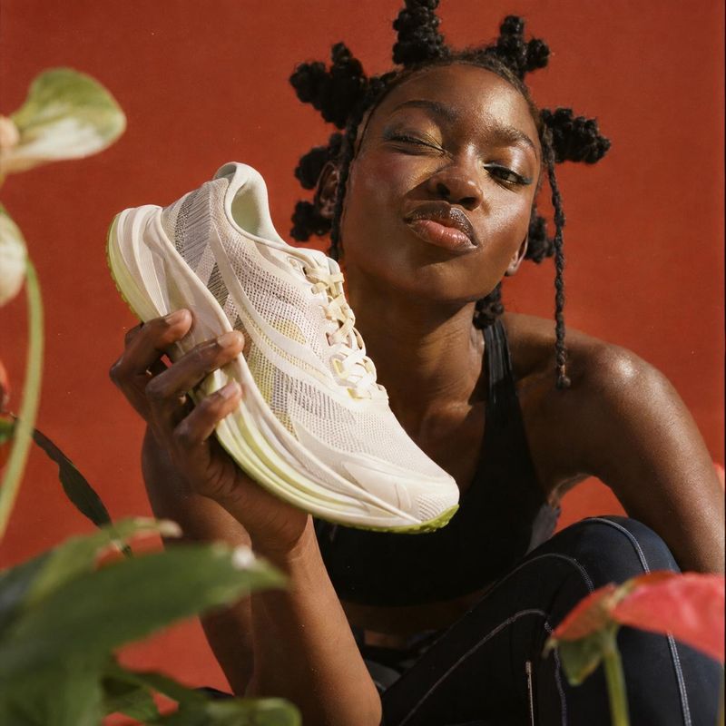 running shoe product in hand product photo: A woman in black athletic wear holds a cream-colored running shoe close to her face against a warm rust-red studio…