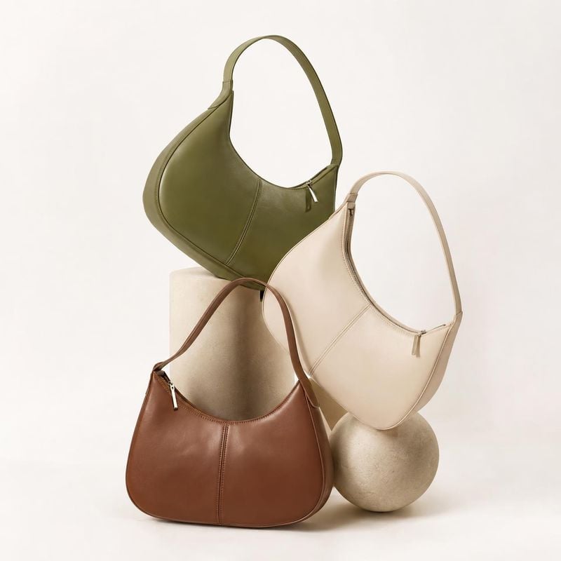 handbags group lineup product photo: Three sculptural leather shoulder bags in olive, cream, and brown are artfully arranged on pale stone forms against a warm…